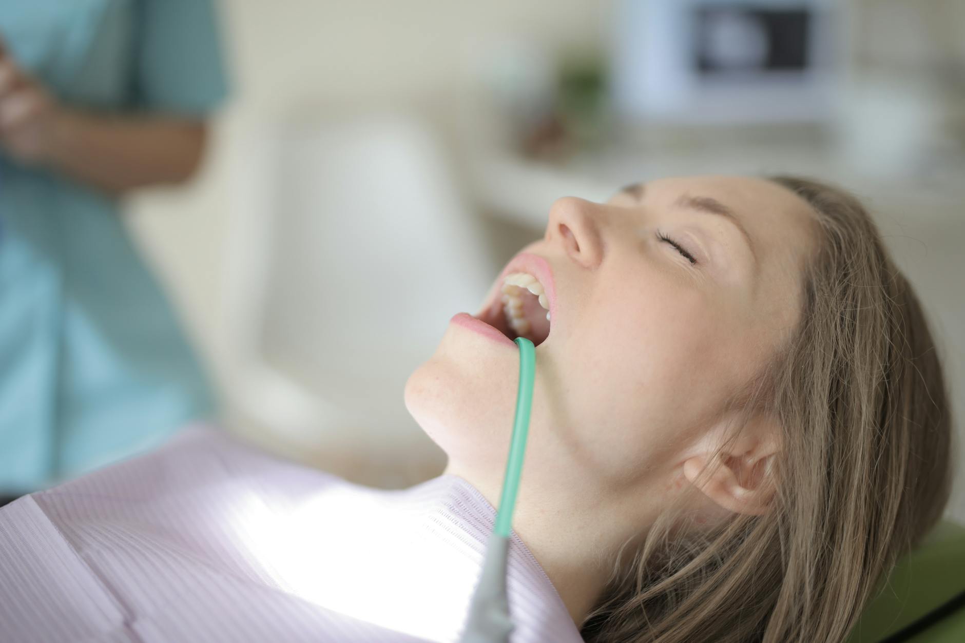 Side view of young female patient with saliva ejector in opened mouth sitting in dental chair while preparing for treatment in modern dental clinic — Atrium Scientific Group